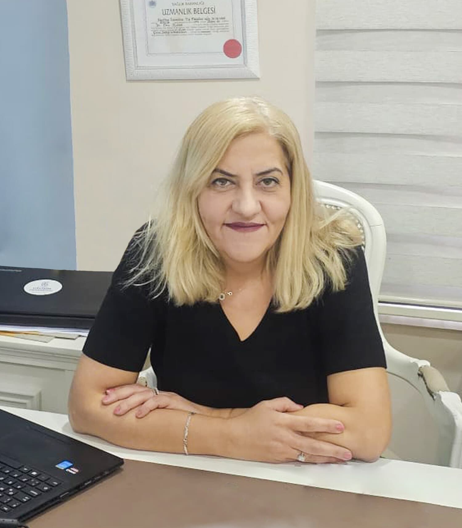 Prof. Dr. Ebru Kolsal muayenehanesinde masasında, arkada uzmanlık belgesi